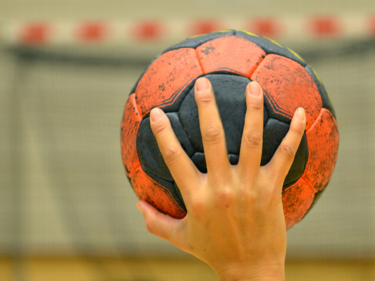 Up close photo of a person holding a black and orange handball with one had about to throw it into a net.