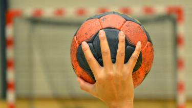 Up close photo of a person holding a black and orange handball with one had about to throw it into a net.