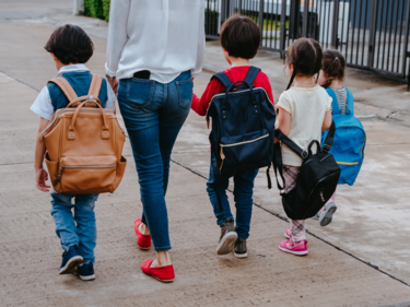 Photo of a parent walking away with four children wearing backpacks, all holding hands. 