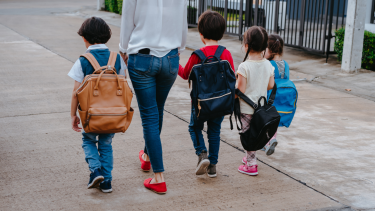Photo of a parent walking away with four children wearing backpacks, all holding hands. 