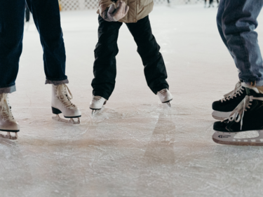 A photo of three people wearing skates standing on ice. Photo is from the knees down.