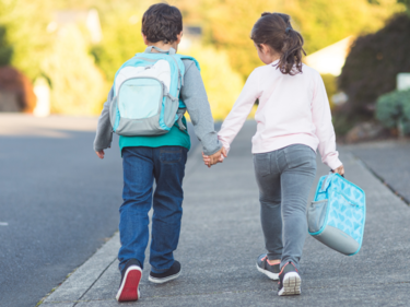 Photo of two children holding hands, a boy and a girl, walking away from the camera. 