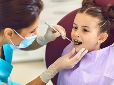 A photo of a girl at the dentist. A hygienist is checking her teeth with a small mirror tool. 