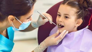 A photo of a girl at the dentist. A hygienist is checking her teeth with a small mirror tool. 