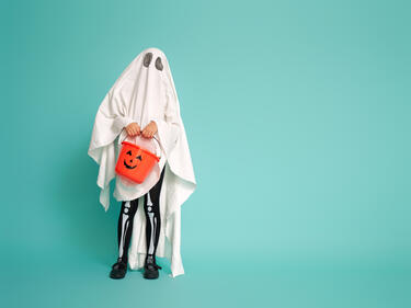 Image of a child dressed as a ghost, holding a pumpkin candy bucket against a blue backdrop.